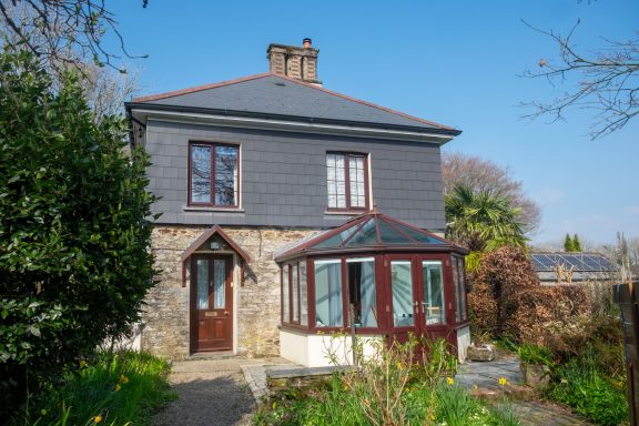 The front of the house, facing South A two-storey house with a stone front and a glass conservatory surrounded by greenery.