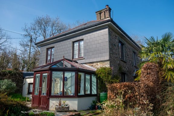 The front of the house, facing South Detached house with a conservatory, surrounded by greenery and clear blue sky.