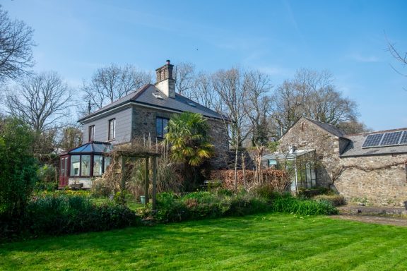 The house as seen from part of the extensive gardens A stone house with a garden, conservatory, and trees under a clear blue sky.