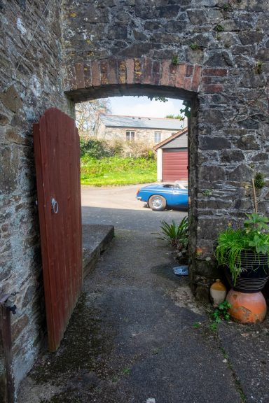 Archway leading to the tarmac back yard area and a double garage View through a stone archway with a wooden door, leading to a street and parked car.